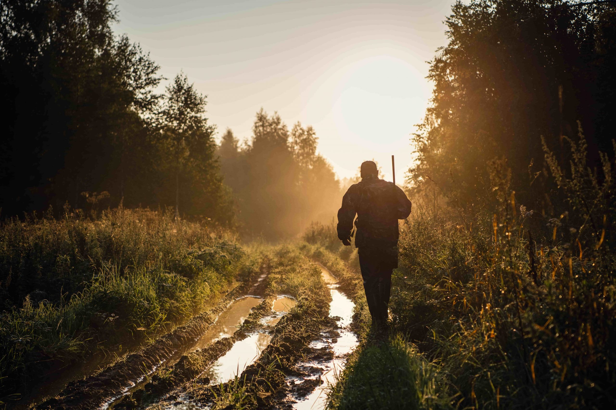 A hunter walks toward a sunset on a muddy forest path