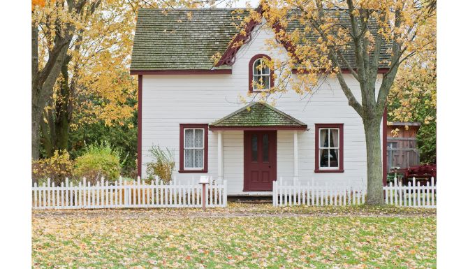 1.5 story white house with red trim white a white picket fence and fall leaves on the lawn