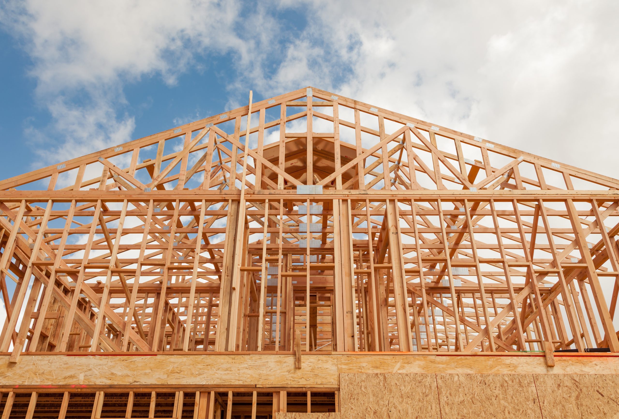 A ground photo pointed towards the sky of a wooden building frame with a blue sky with white clouds.