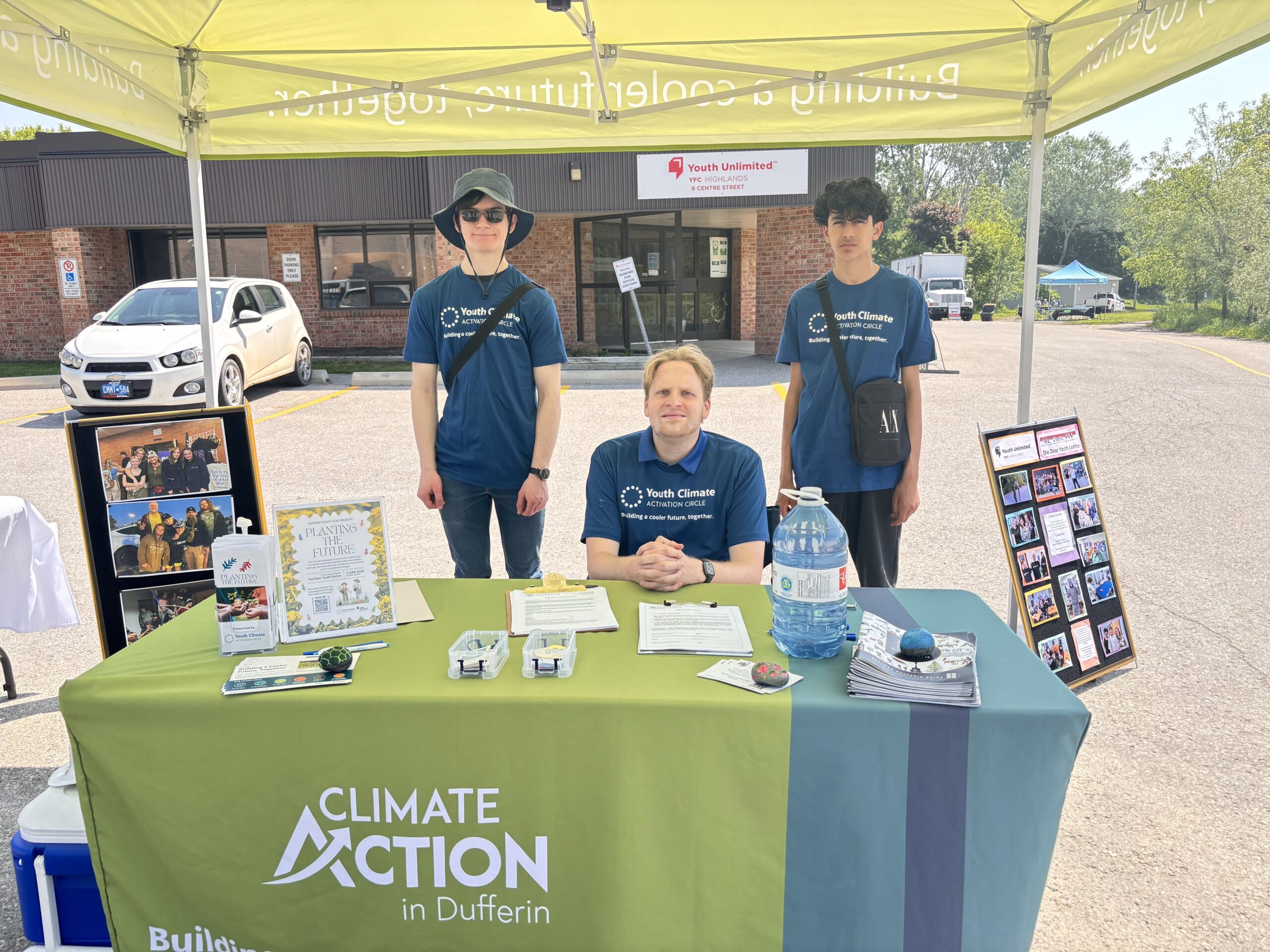 Three youth climate ambassadors stand behind a Climate Action in Dufferin table, hosting the Welcome Booth for a native plant gardening event